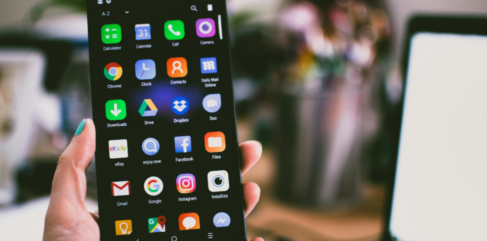 A hand holding a smartphone displaying a colorful home screen with various app icons, including calendar, clock, camera, drive, and social media apps. The background is blurred, showing a desk with office supplies and a computer monitor.