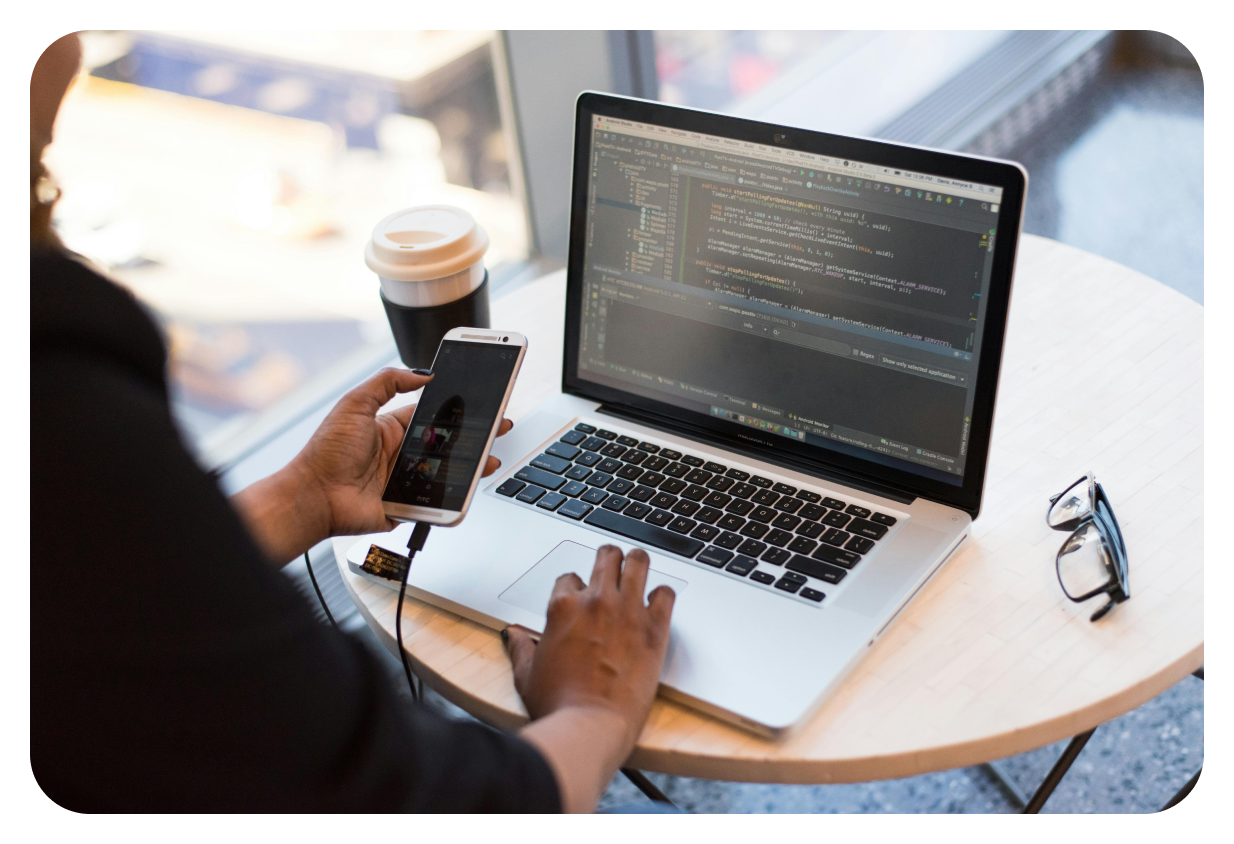 A person sits at a round table with a laptop displaying code, a smartphone, and a coffee cup with a lid. Glasses are set to the side.
